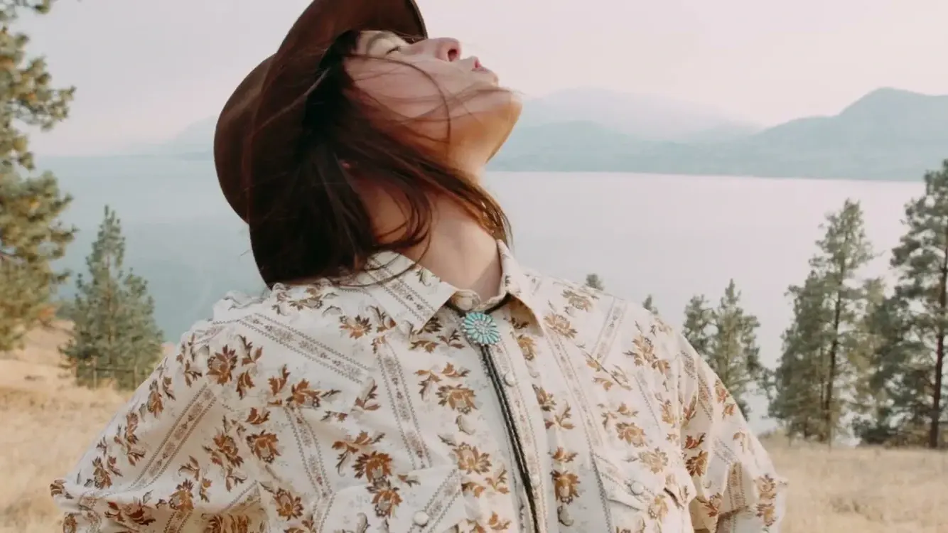 A hatted figure tilts their face upward on a dry hillside above a lake in Autumn Sun.