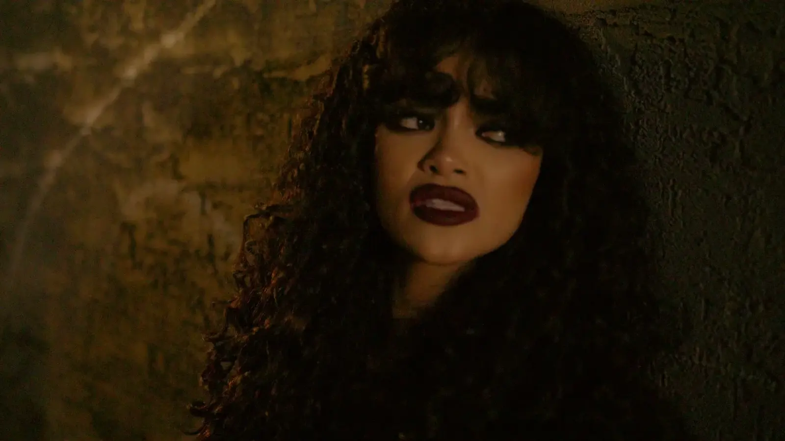 A woman sings beside a rough wall in warm light.