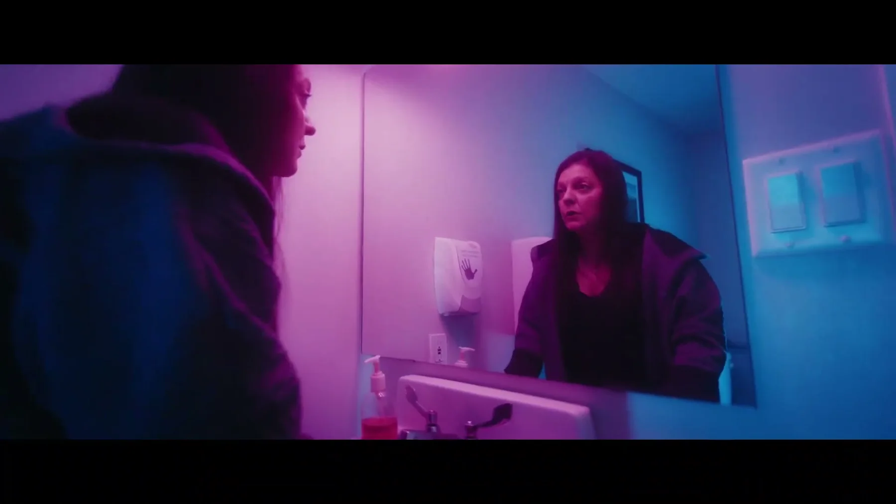 A woman faces her reflection in a magenta and blue bathroom mirror.